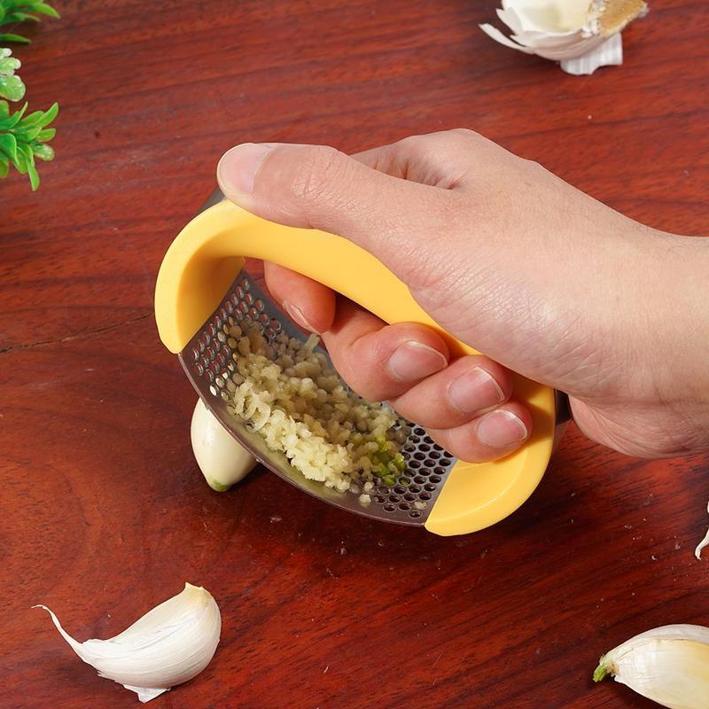 Hand using a yellow garlic press on a wooden surface with garlic cloves around.